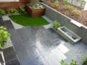 Black limestone patio with artificial turf on top left corner. Stainless steel weir waterfall on right back of patio.