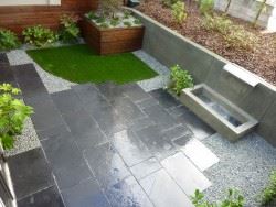 Black limestone patio with artificial turf on top left corner. Stainless steel weir waterfall on right back of patio.