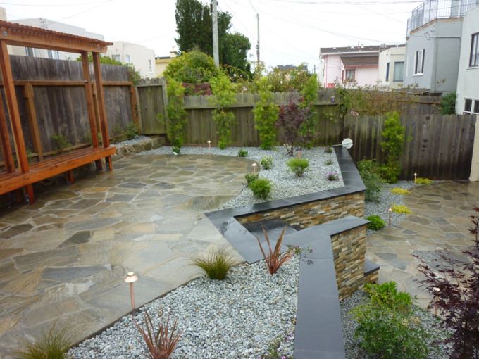 Elevated quartzite flagstone patio on left on top of retaining wall in Lakeshore, San Francisco.  CMU retaining wall veneered with stone in different shades of brown. Wood sitting area in far left of patio with pergola.  Various plants surround patio with crushed rock.  Below the patio is a quartzite walkway on the right side. Plants surrounding pathway.  