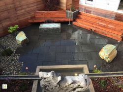 Black basalt patio in middle with redwood benches in back and back right. Fire pit with blue rocks in front of back bench. One turquoise boulder on left of fire pit and another on right of bench. River rock on left of patio.