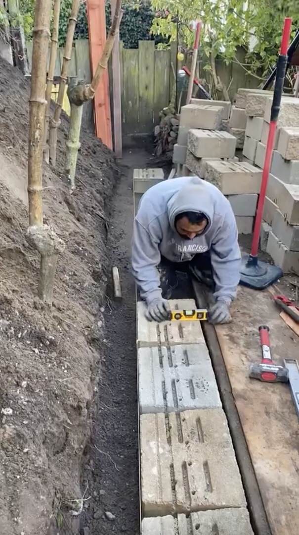 Worker in light blue hooded sweatshirt on knees looking at level positioned on top of Versa-Lok base block. Versa-Lok blocks stacked in right background. To left is dirt hillside.