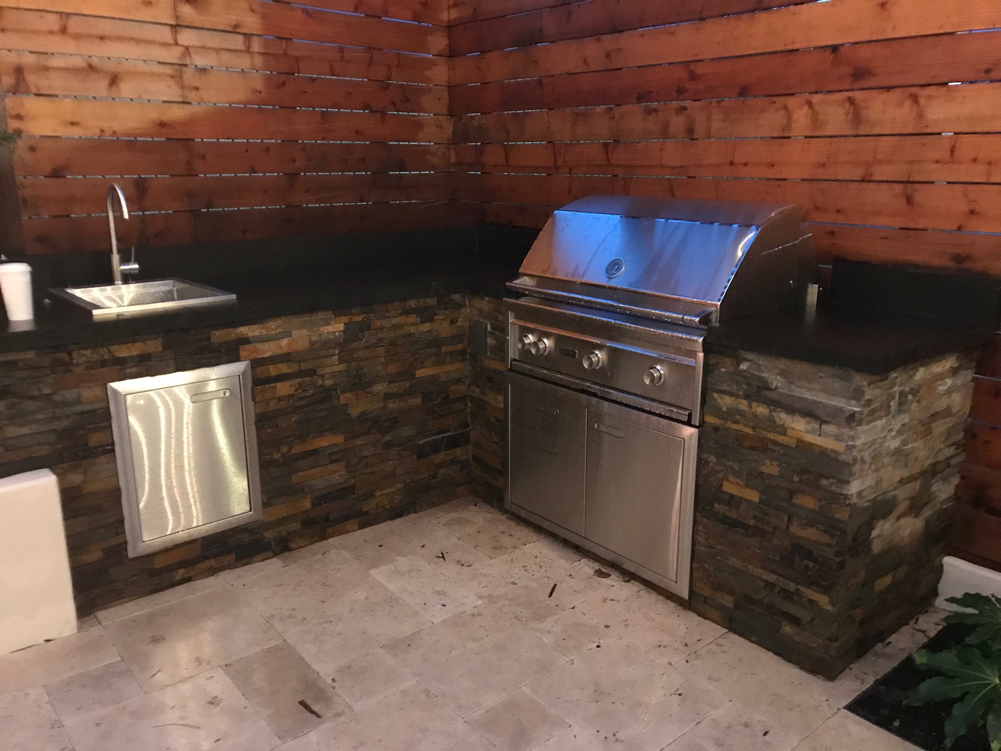 Custom built in BBQ unit on right with sink on left and stone counter in L-shape. Redwood fence in background and travertine pavers on ground.
