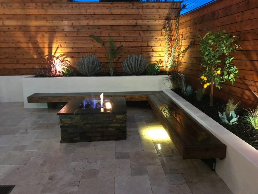 Fire pit in foreground, raised planters in back and right side with L-shaped redwood seating. Redwood fence in back of planters and travertine pavers on ground.