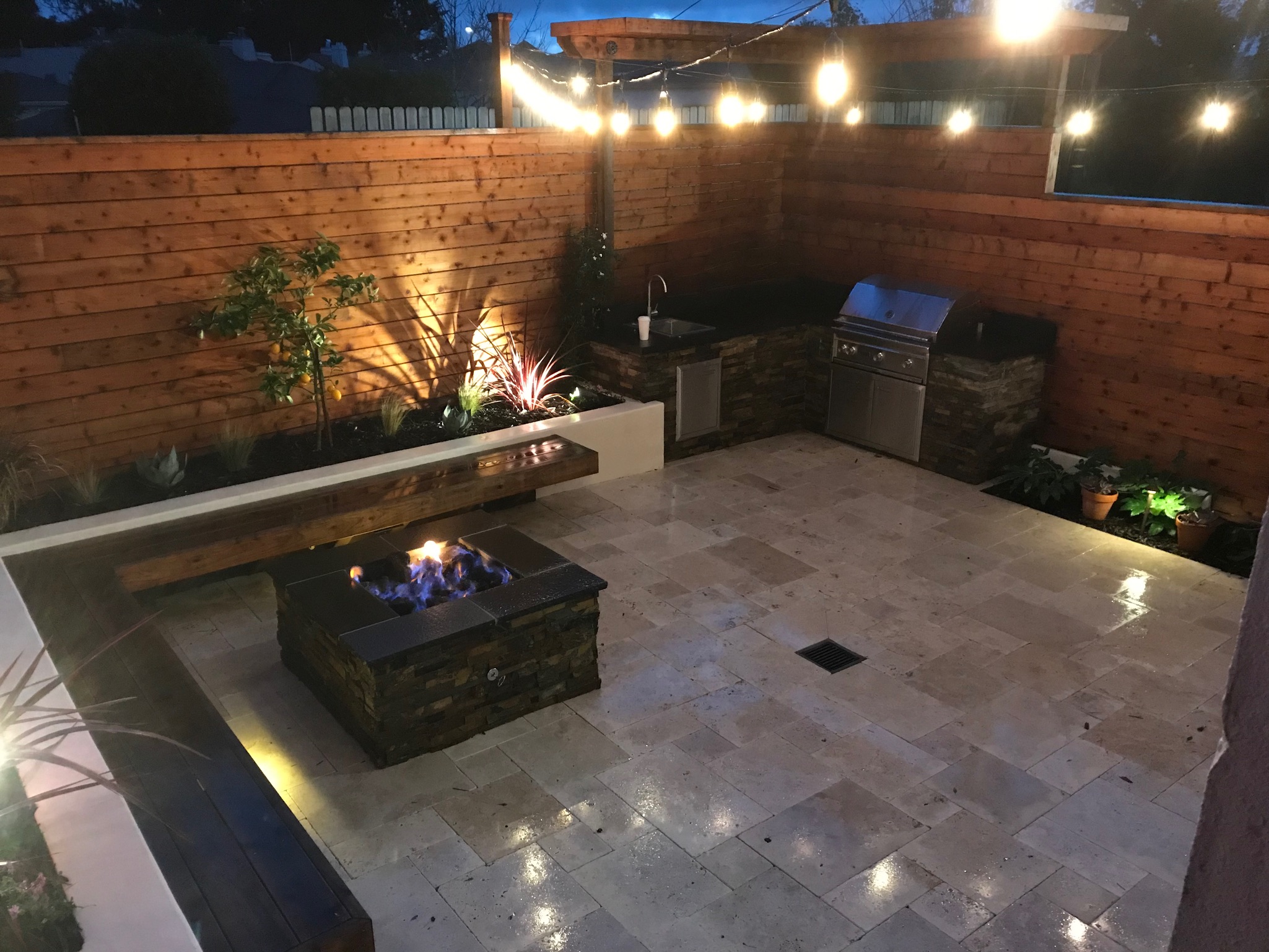 Modern, sustainable backyard complete with BBQ area and sink in back right corner. Fire pit and L-shaped redwood seating, backed with raised planters in left corner. Travertine pavers on ground and redwood fence in background.