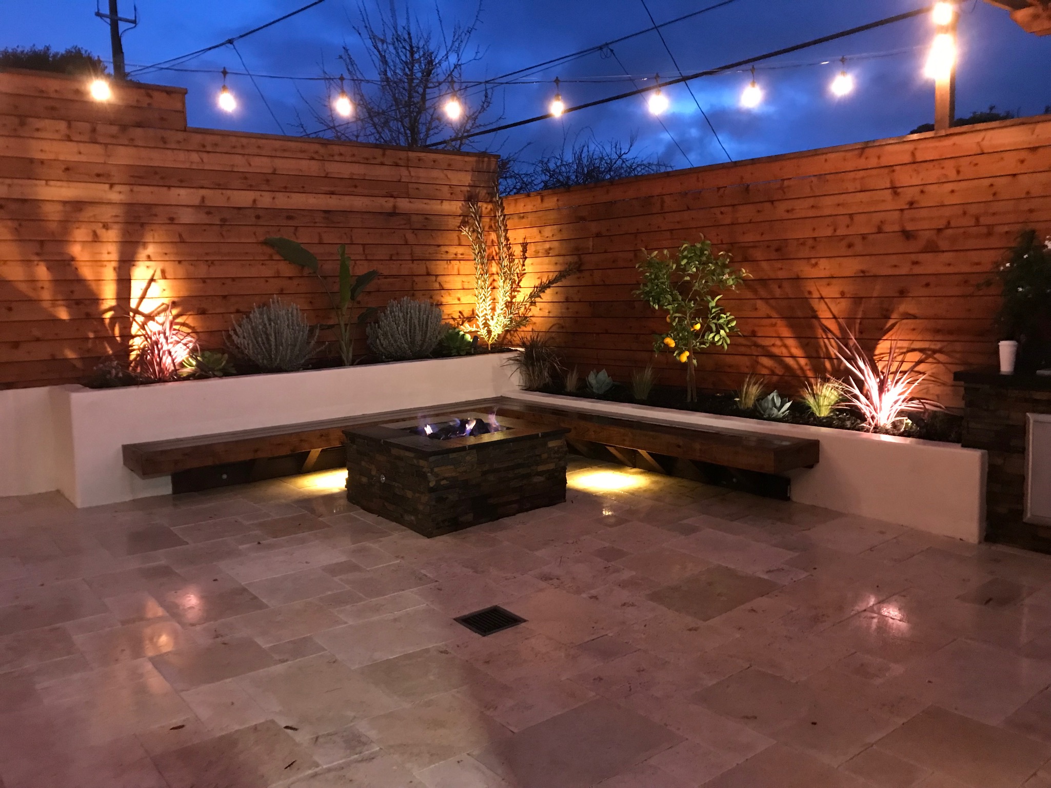 Fire pit in foreground, raised planters in back and right side with L-shaped redwood seating. Redwood fence in back of planters and travertine pavers on ground. Stringer lights above.