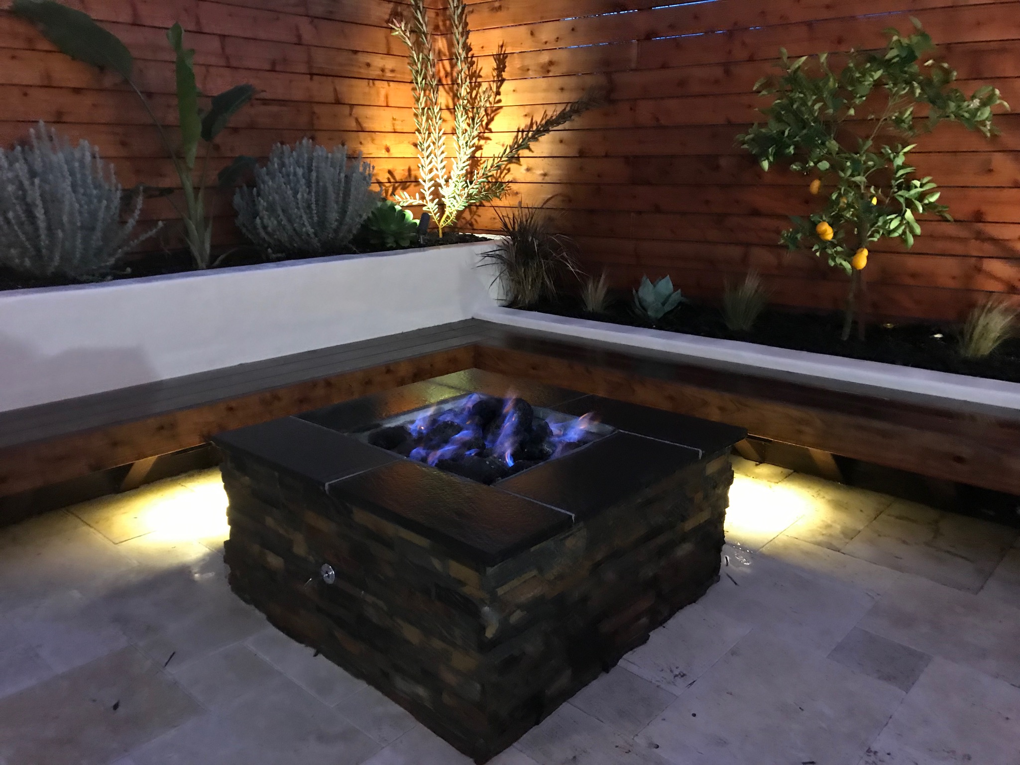 Close-up of lit fire pit in foreground with blue flames. Raised planters, plants, and redwood fence in background.