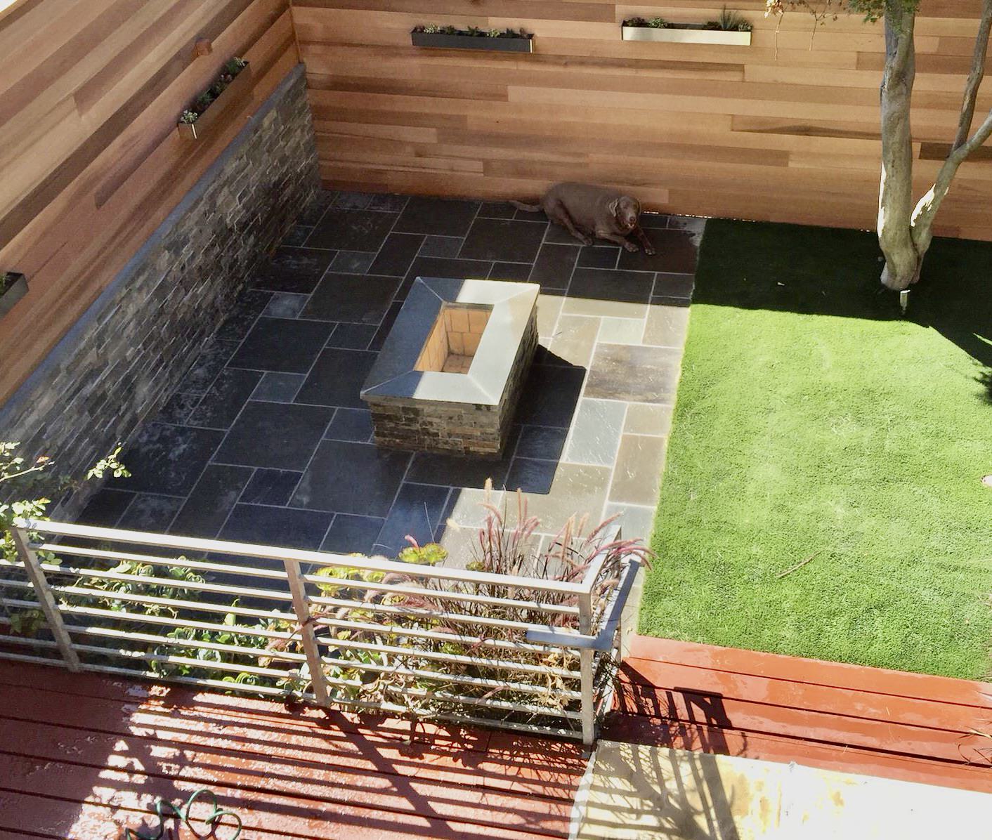 Bluestone patio on back left with fire pit in center.   Artificial turf to right of patio. Cedar fence surrounding the landscape on left and back of patio and artificial turf.