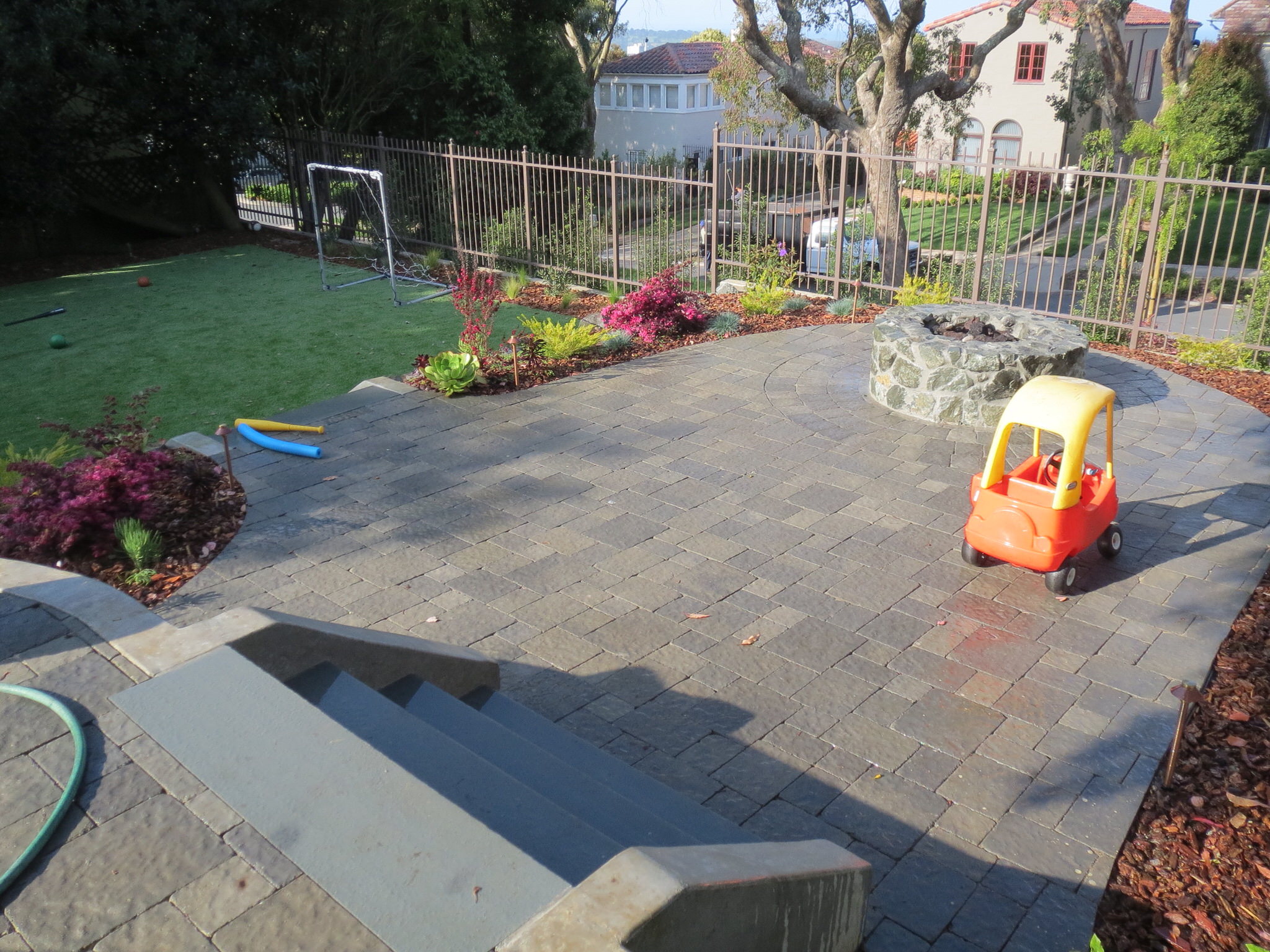 Gray interlocking paver patio in foreground.  Red and orange child's car toy on the right of patio and round fire pit on left.  Artificial grass on far left back side.  Various plants surround patio with bark.