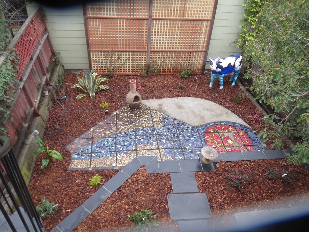 Free-form mosaic-designed tile patio. Cow statue in right back. Patio surrounded by bark and various plants.