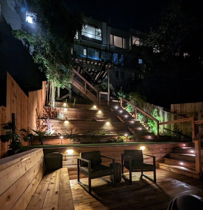 View of a terraced back yard in the Bay Area. Outdoor lighting highlights stairs, plants, and bottom deck. The patio can be seen from the bottom deck.