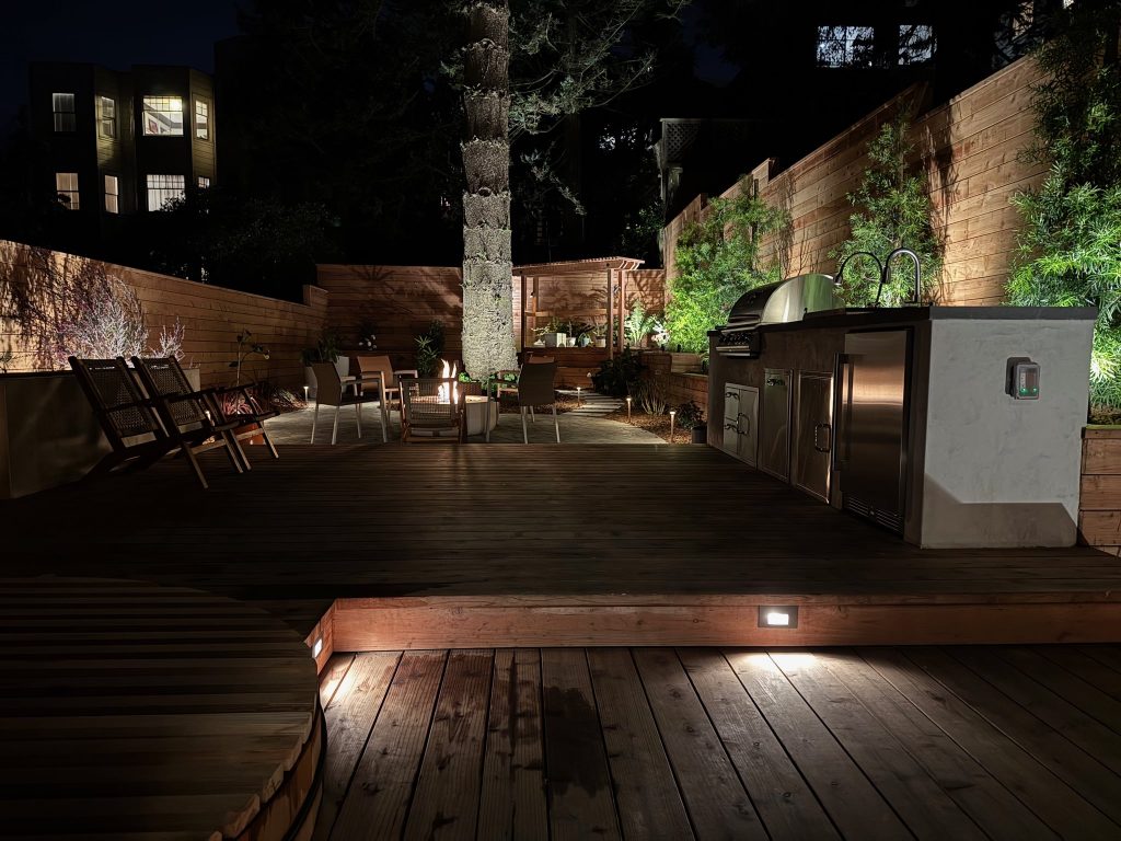 Completed backyard at night with redwood deck and outdoor kitchen in foreground.
