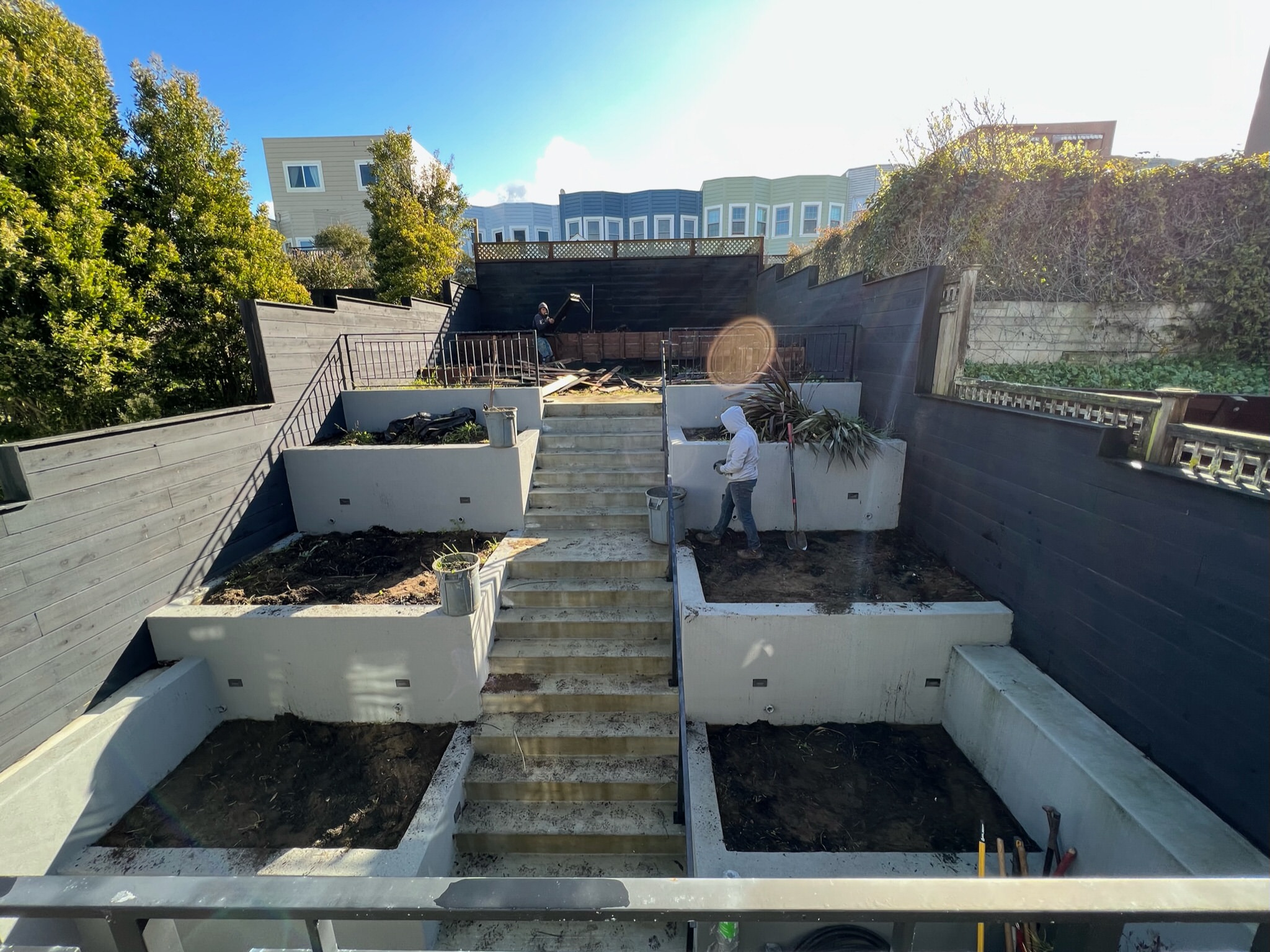 Backyard before at our Outer Sunset, San Francisco project. Three tiered retaining walls filled with dirt and some dying plants on either side of stairway leading up to top level patio. Top patio area covered with debris of wood and dirt.