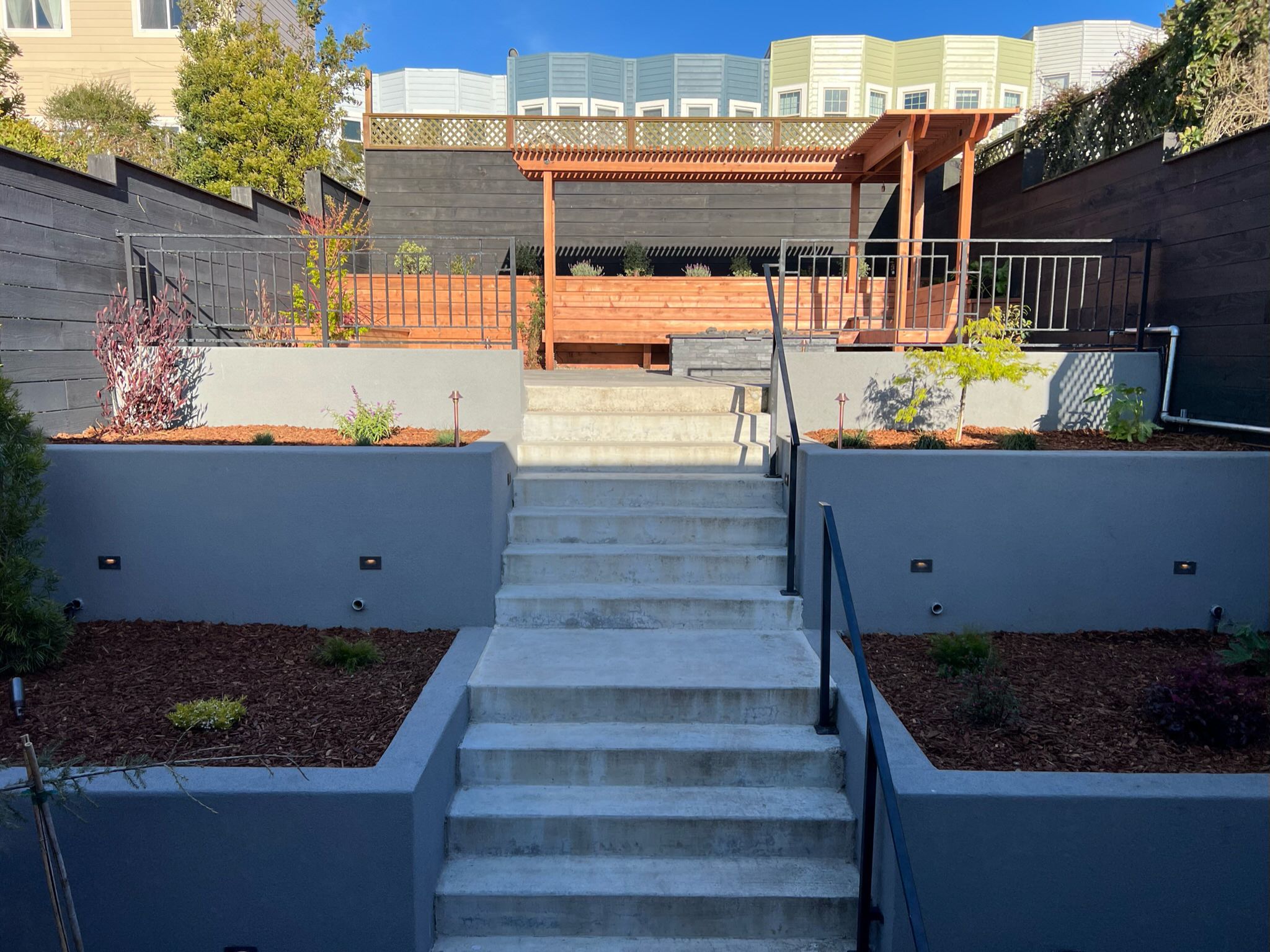 Completed backyard at our Outer Sunset, San Francisco project. Two tiered retaining walls on right and left side of stairway leading up to top level patio area. Two tiered retaining walls with plants. Top tier patio area with redwood bench and pergola above.