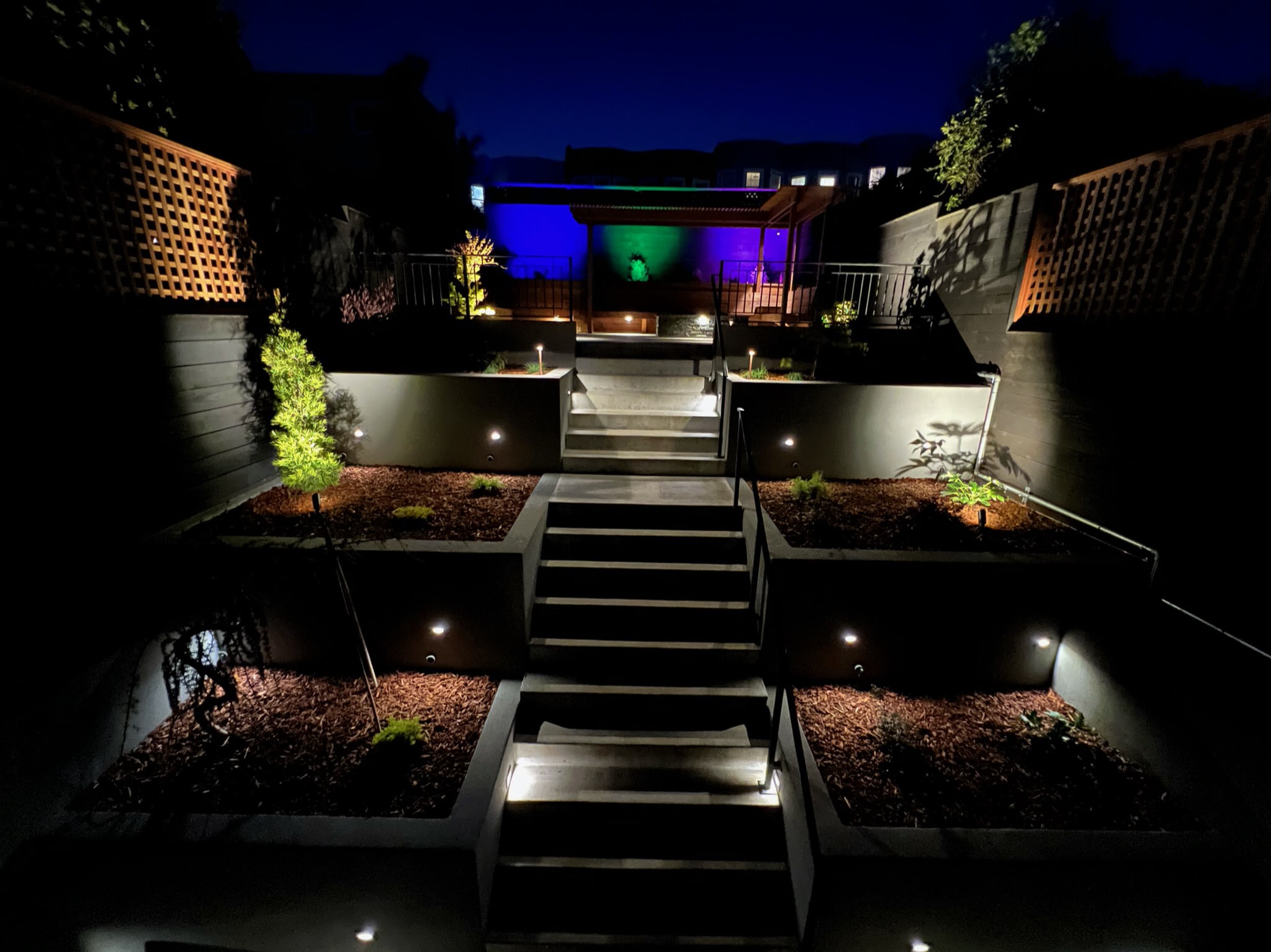 Completed backyard at night at our Outer Sunset, San Francisco project. Three tiered retaining walls on right and left side of stairway leading up to top level patio area. Retaining walls are dimly lit up with plants. Top tier patio area lit up with blue, green, and purple lighting in background behind pergola and bench.