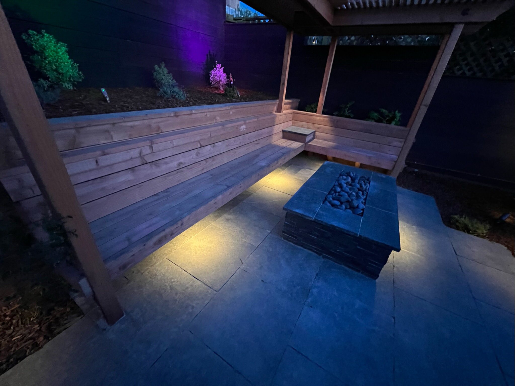 Redwood bench, pergola, fire pit, and basalt patio at night at our Outer Sunset, San Francisco project. Warm yellow lights under bench. Purple and green lights in back of raised planted behind bench.