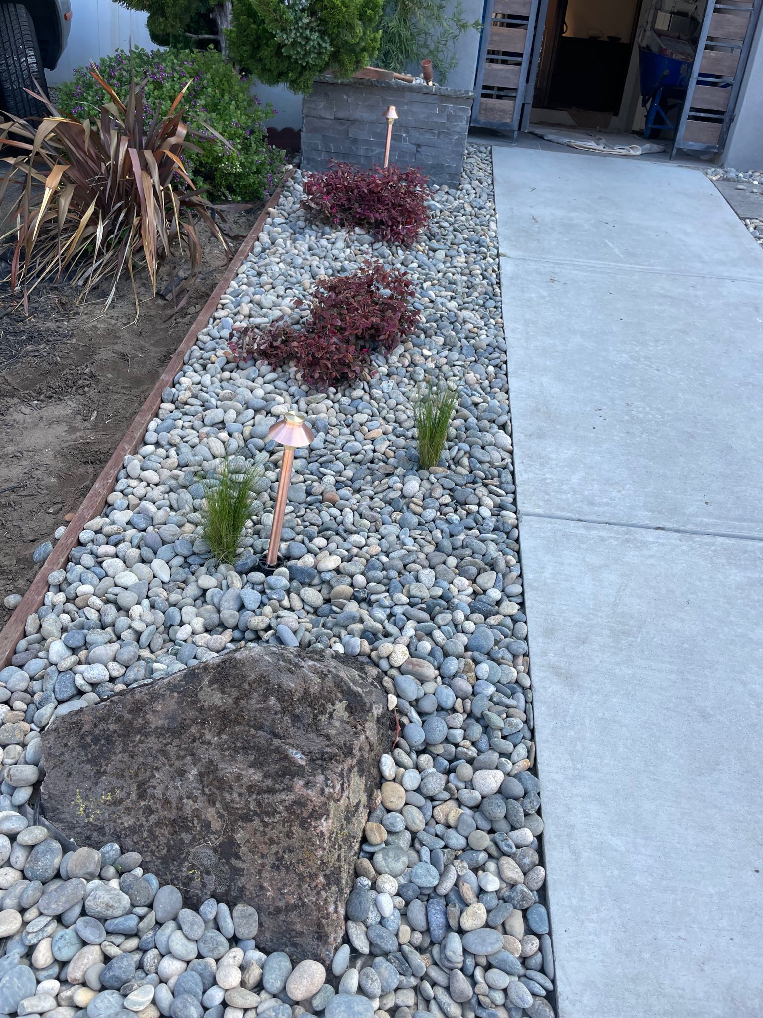 Completed front yard at Outer Sunset, San Francisco project. Boulder in front and raised planter in back. Two Chinese fringe plants planted next to each other vertically. Two Mexican Feather grass plants in back of boulder and two copper light fixtures. Ground covered with Lynn Creek stones.