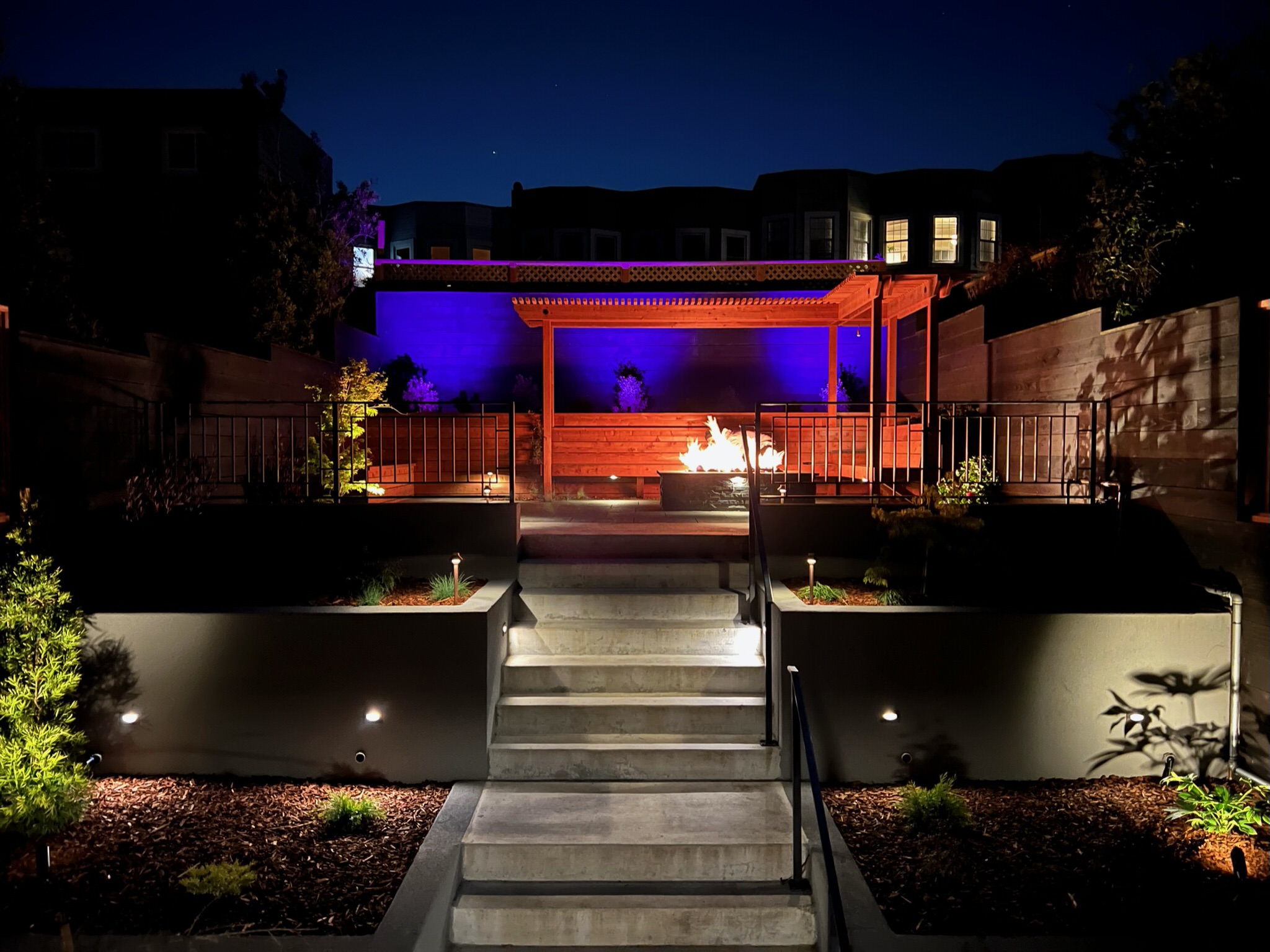 Backyard at night with FX Luminaire Luxor lighting system. Purple background color behind pergola. Fire pit lit with flames visible in front of redwood bench and pergola.