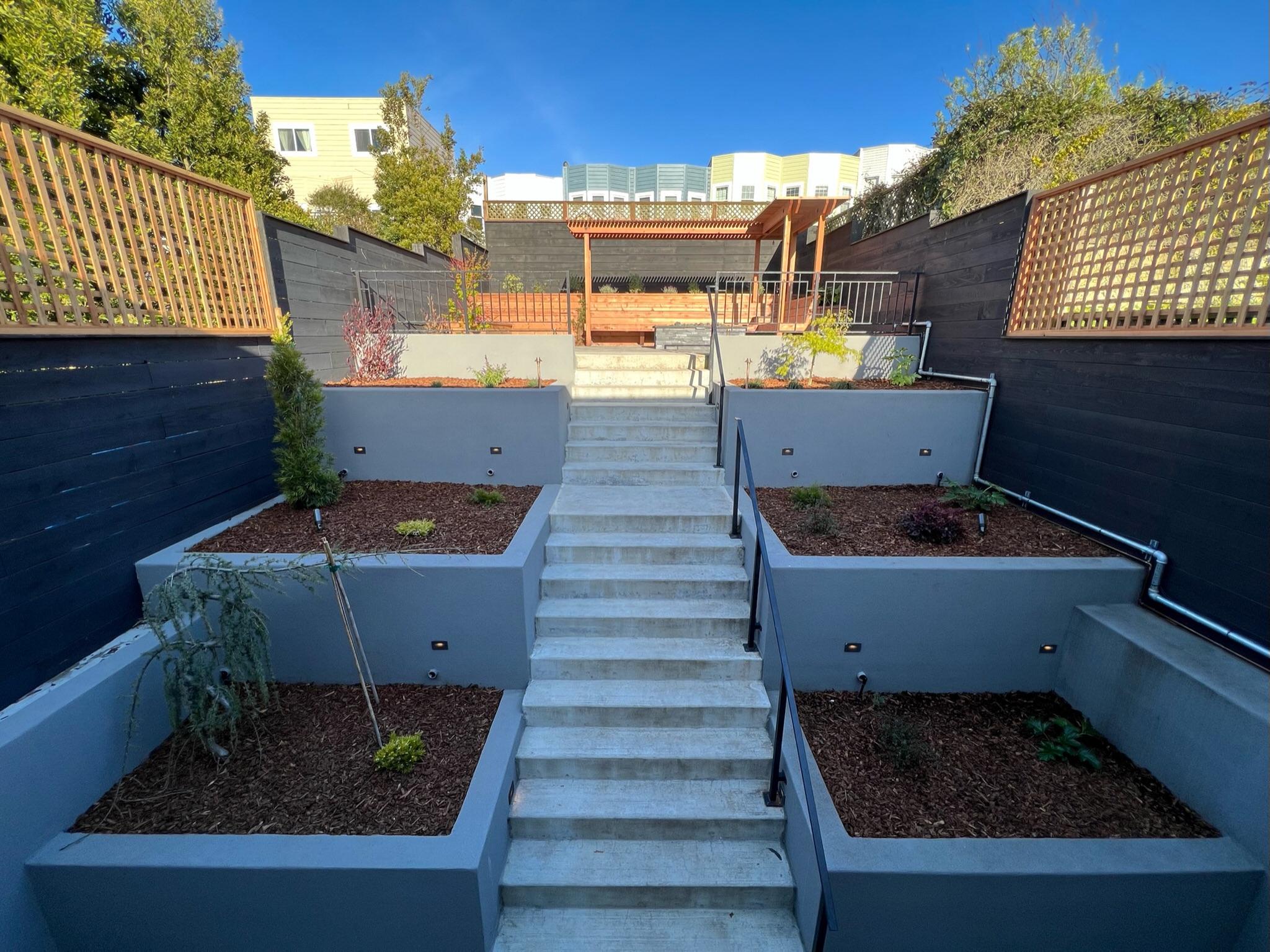 Completed backyard at our Outer Sunset, San Francisco project. Two tiered retaining walls on right and left side of stairway leading up to top level patio area. Two tiered retaining walls with plants. Top tier patio area with redwood bench and pergola above.