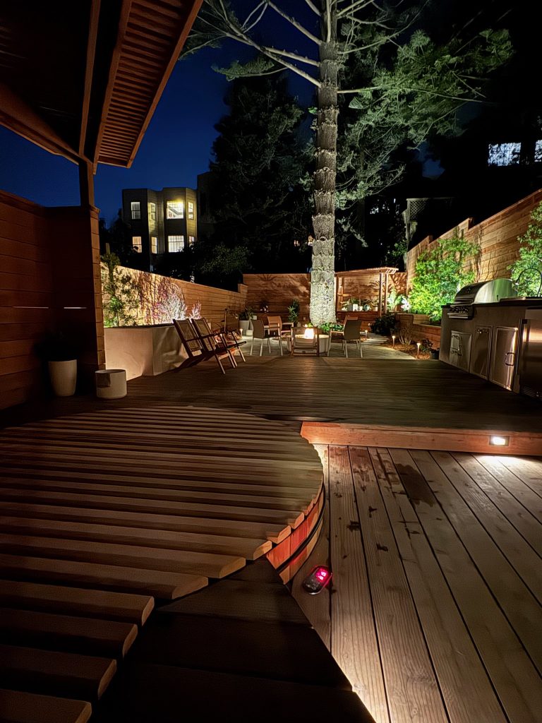 Completed backyard at night with covered hot tub in the foreground. Uplights highlight Norfolk pine in the back middle of the landscape. Deck lighting and raised planters around the perimeter of the landscape also lighted.